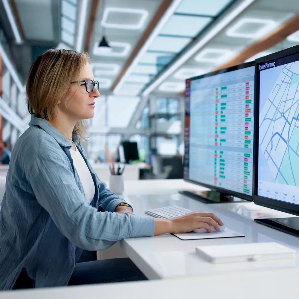 Woman wearing glasses working on a computer with dual monitors displaying data and a route planning map in a modern office.