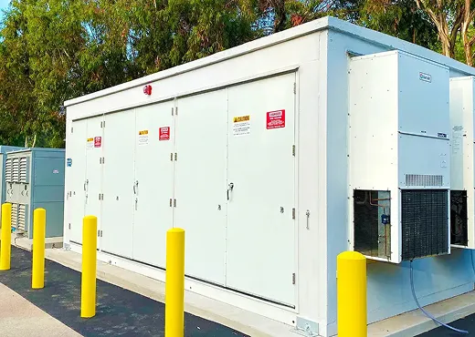 Large white outdoor battery energy storage unit with yellow bollards and surrounded by trees.