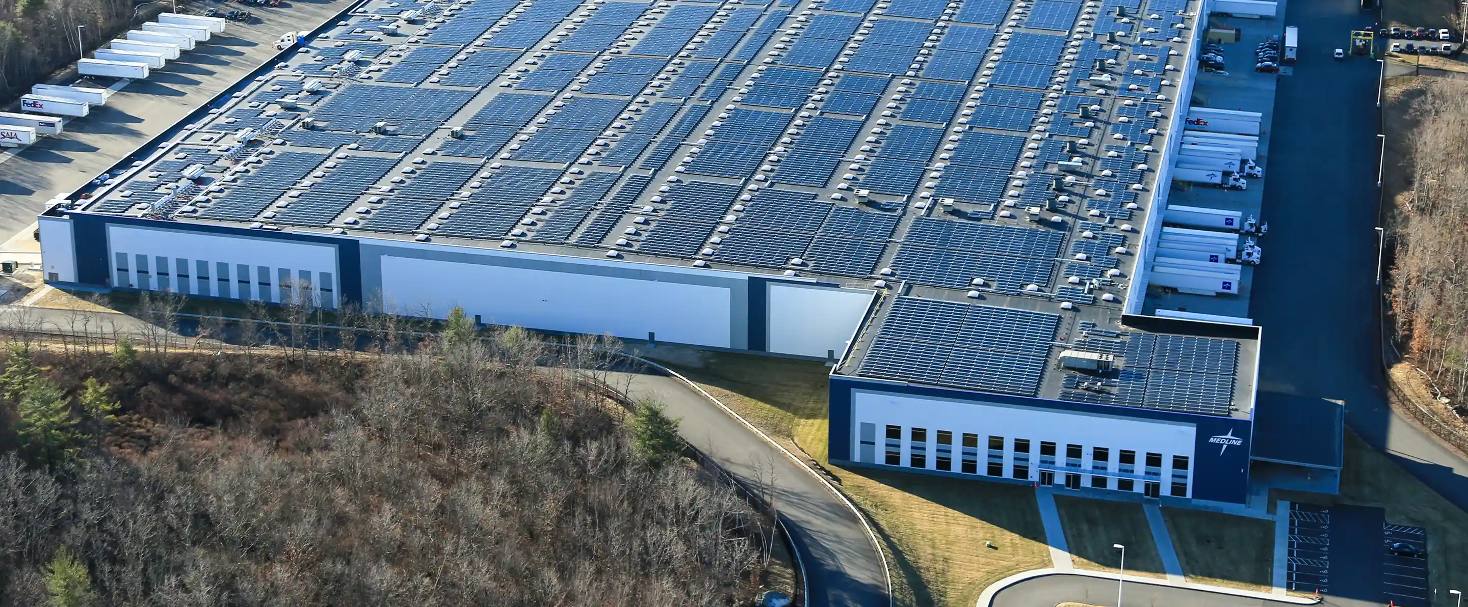 A large industrial warehouse with solar panels covering its roof and trucks parked at loading docks.