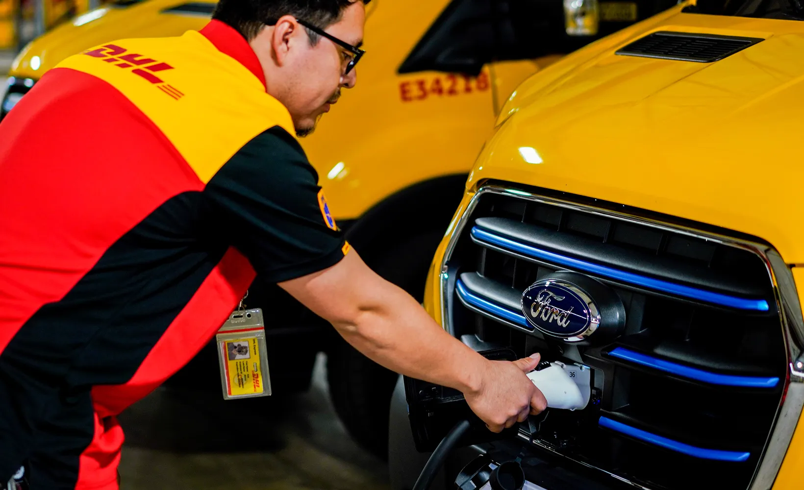Man wearing a DHL uniform charging a yellow Ford electric vehicle.