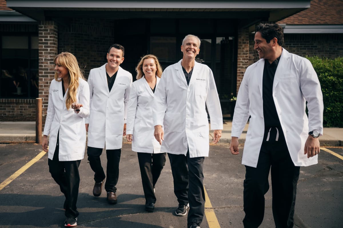 Five dental professionals in white coats walking together outside a medical building.