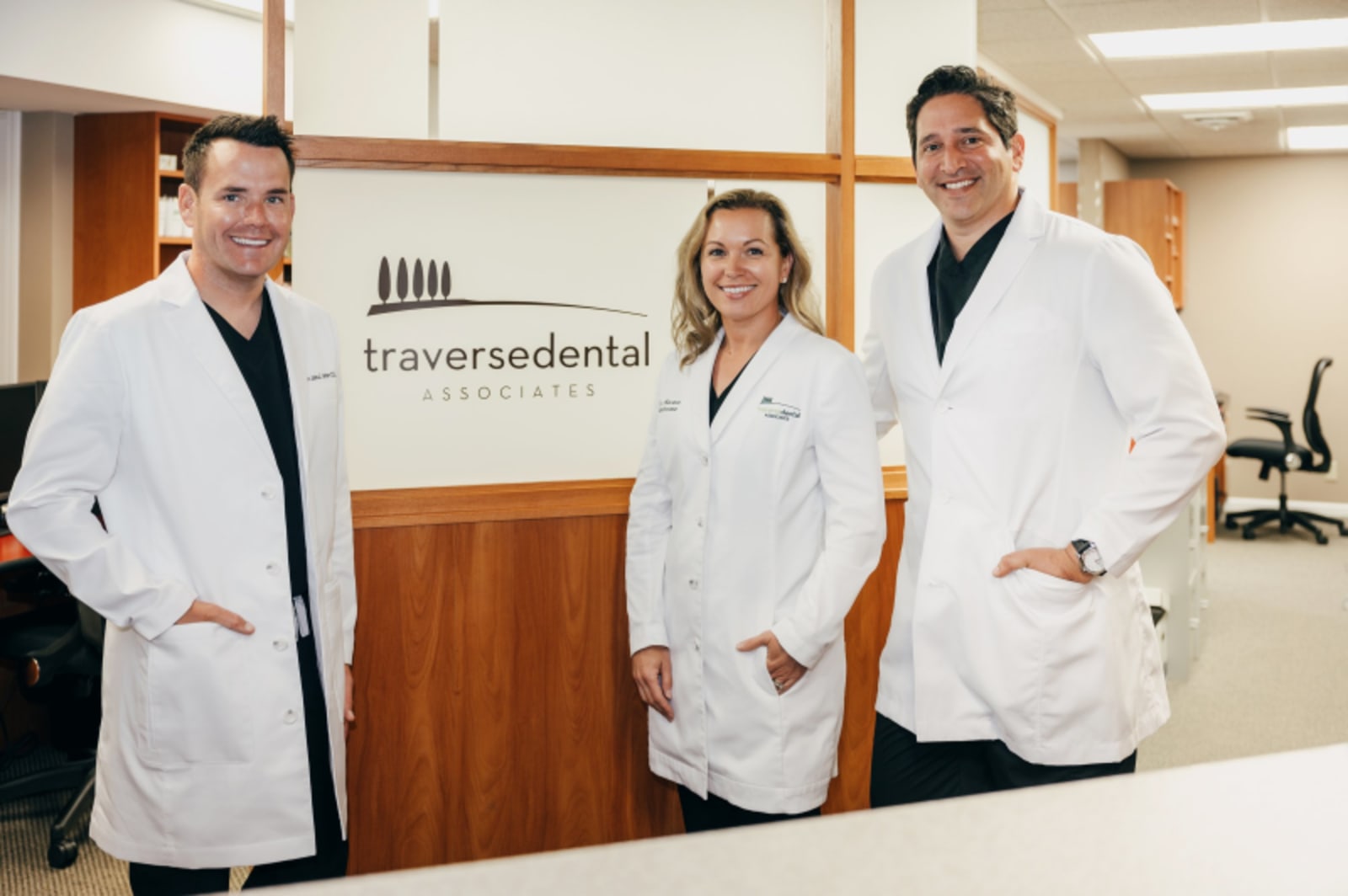 Three dental professionals in white coats smiling at reception desk area