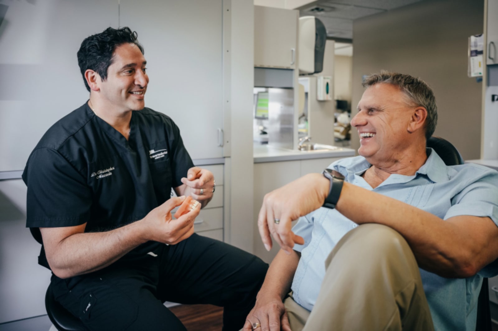 Dentist in black scrubs consulting with male patient in clinical treatment room setting