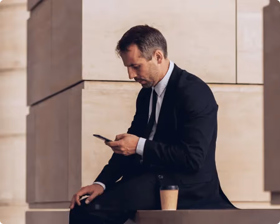 Geschäftsmann in schwarzem Anzug schaut auf sein Smartphone mit Kaffee