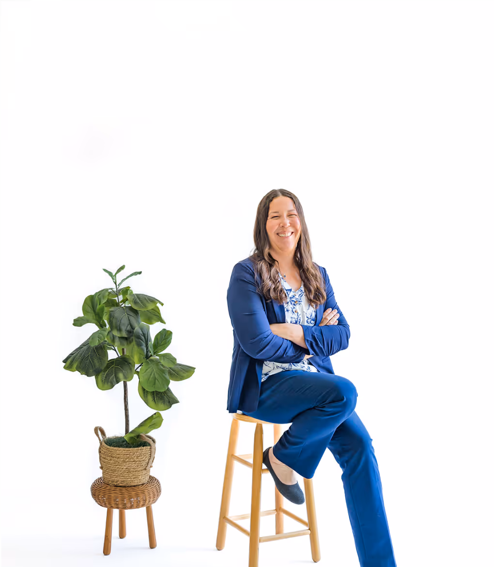 Smiling woman in blue business attire sitting on a wooden stool with a potted green plant on a wicker stool beside her against a white background.