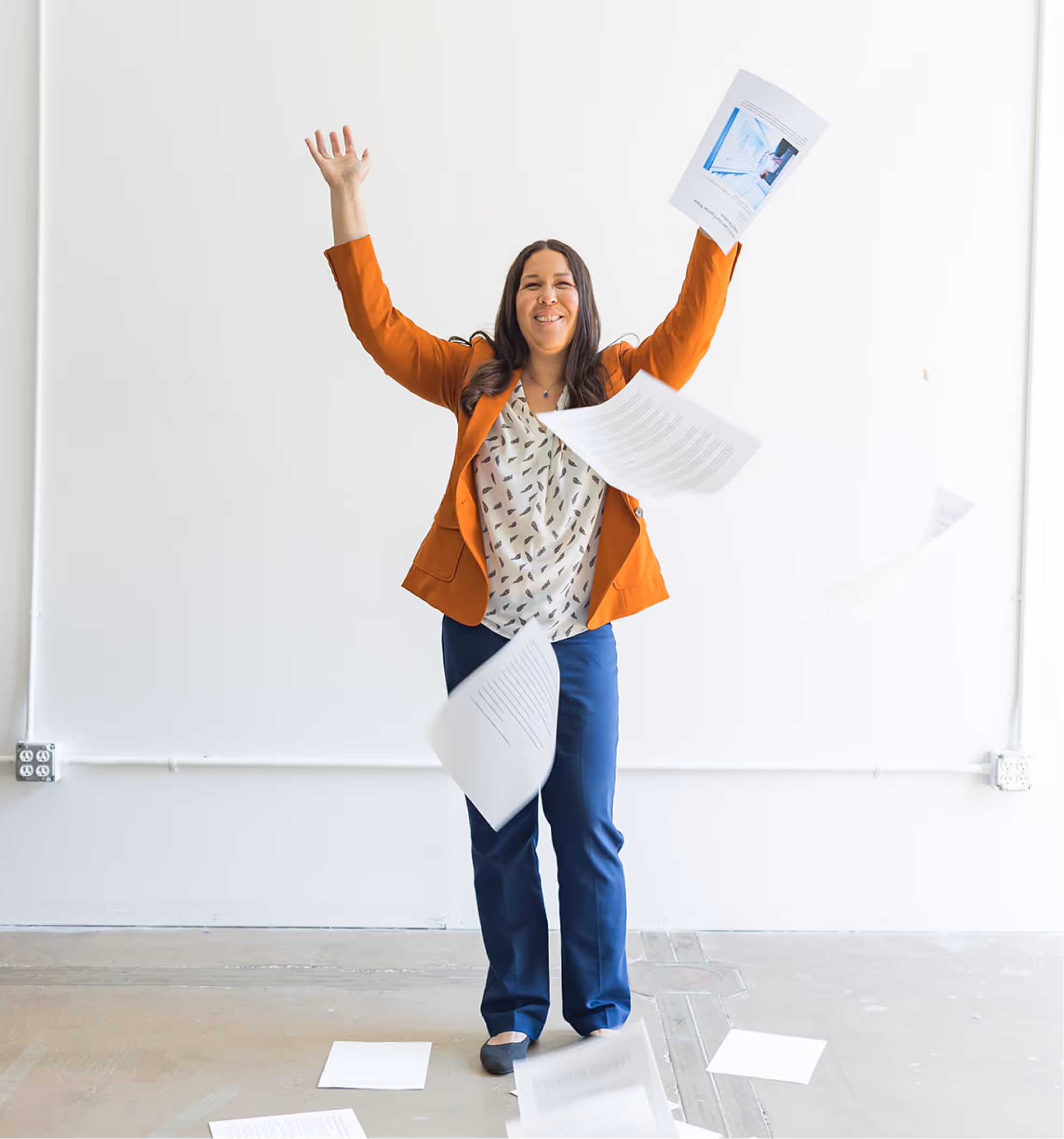 Smiling woman in orange blazer throwing papers into the air against a plain white wall.
