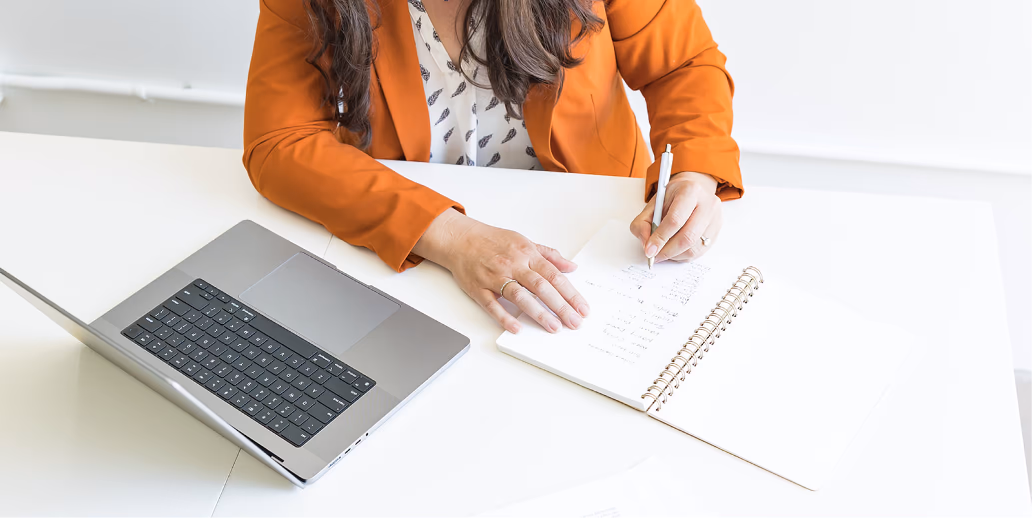 Person in orange blazer writing notes in a spiral notebook next to an open laptop on a white desk.