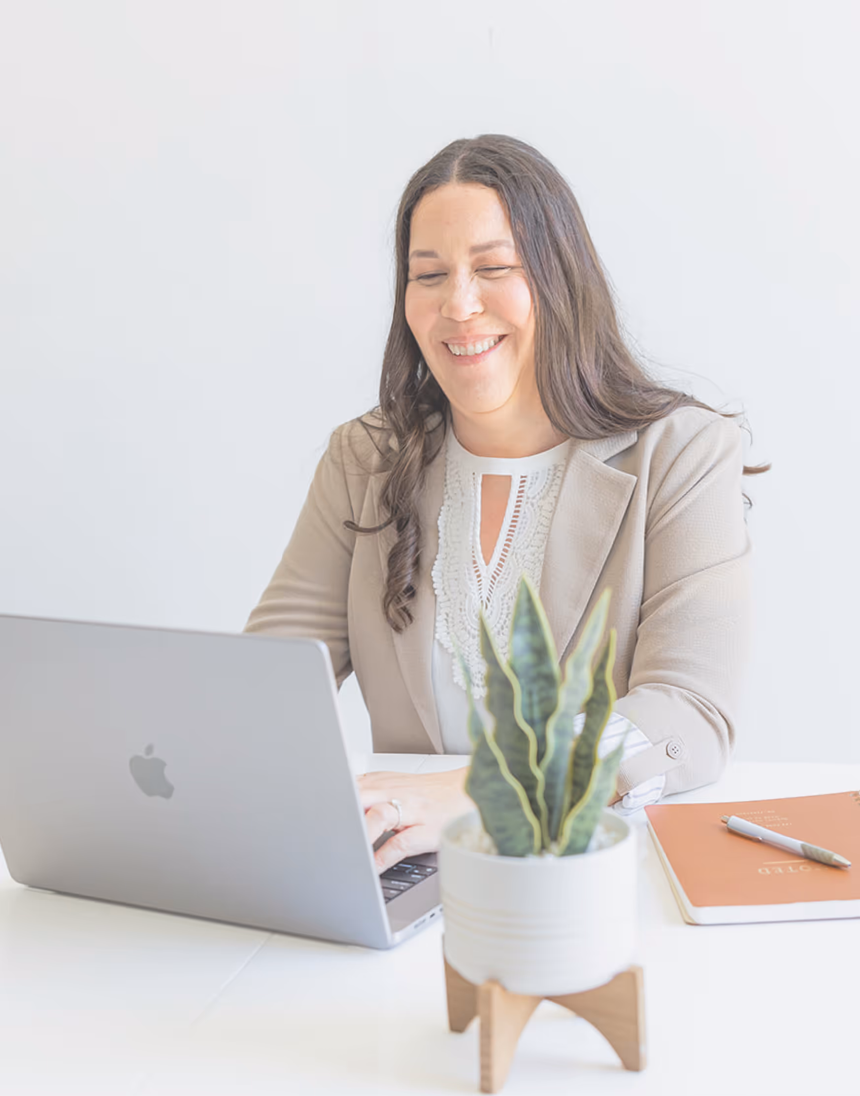 Smiling woman in beige blazer working on a laptop at a white desk with a potted plant and notebook.