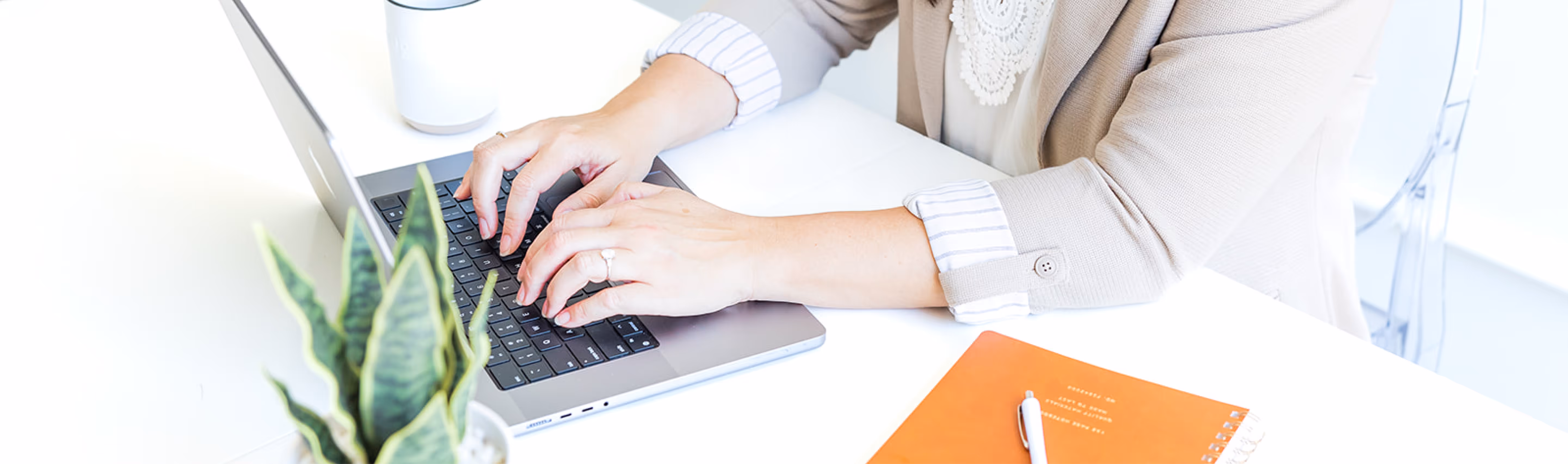Person wearing a beige blazer typing on a laptop at a white desk with a potted snake plant and an orange notebook with a pen.