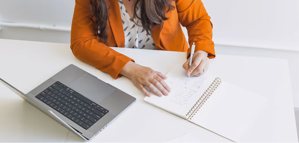 Person in orange blazer writing notes with a pen in a spiral notebook next to an open laptop on a white desk.