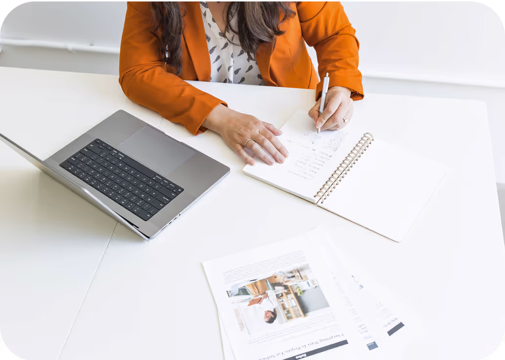 Person in an orange blazer writing notes in a spiral notebook next to an open laptop and printed documents on a white desk.