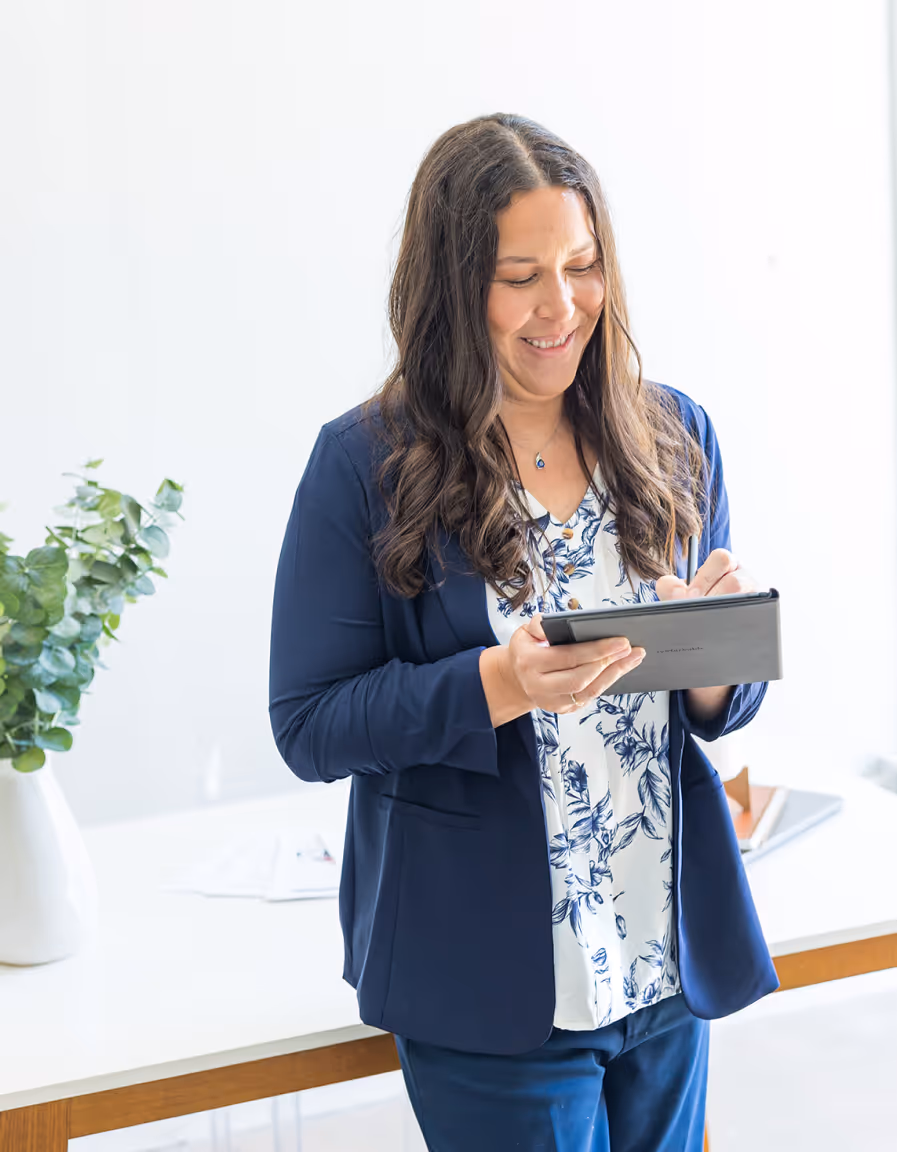Smiling woman in a navy blazer using a digital tablet in a bright office with a plant on the desk.