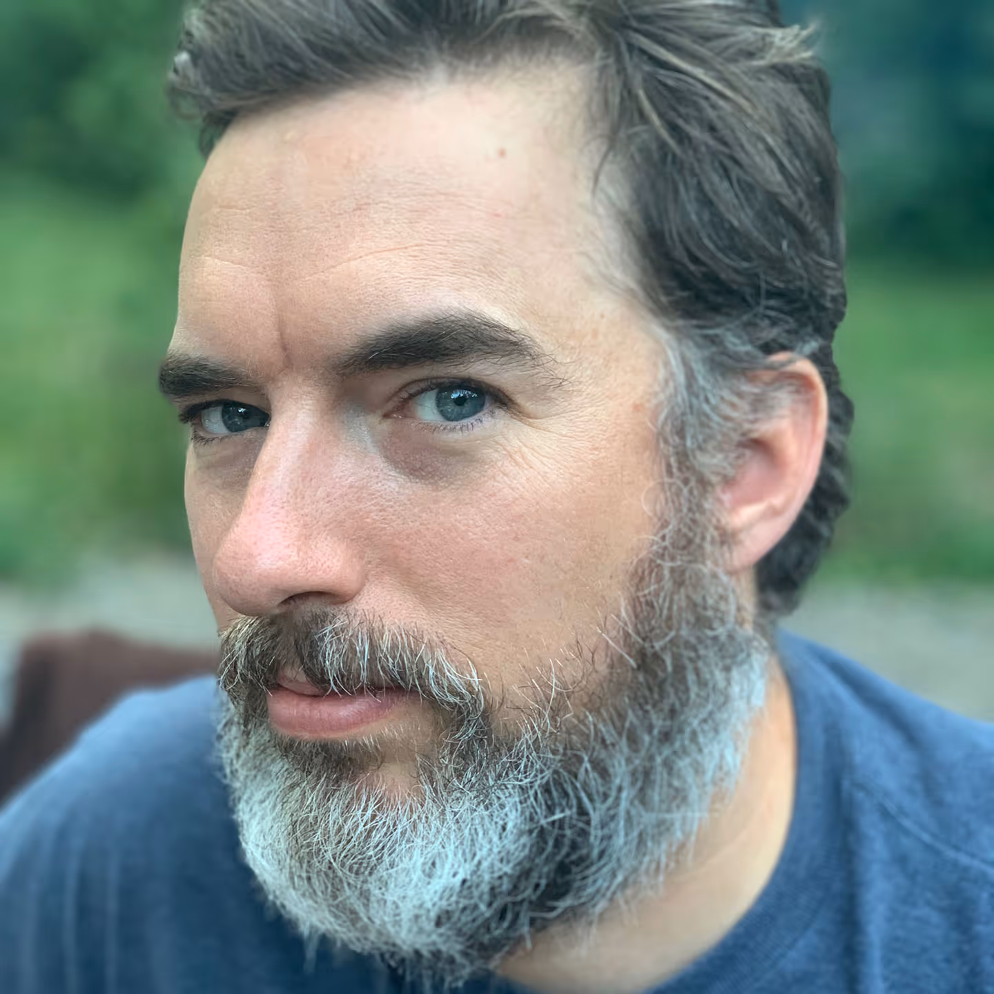 Close-up of a man with blue eyes, dark hair, and a salt-and-pepper beard wearing a blue shirt outdoors.