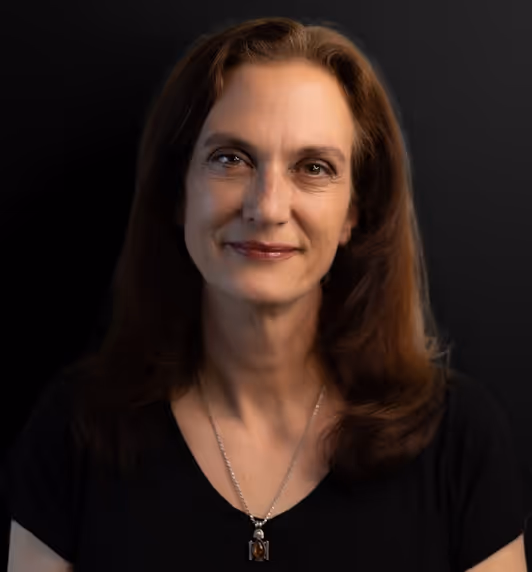 Portrait of a middle-aged woman with brown hair wearing a black top and a pendant necklace, against a dark background.