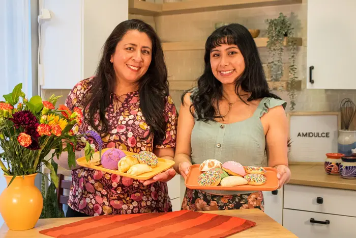 Two women proudly display trays of colorful Mexican pastries, smiling in a bright kitchen filled with flowers.