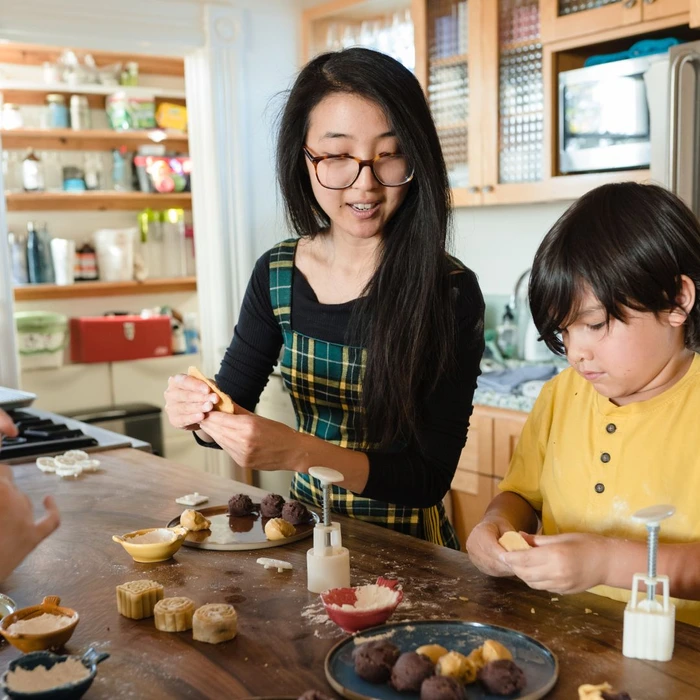 Annie and a child make vegan Asian snacks together in a kitchen, surrounded by ingredients and baking tools.