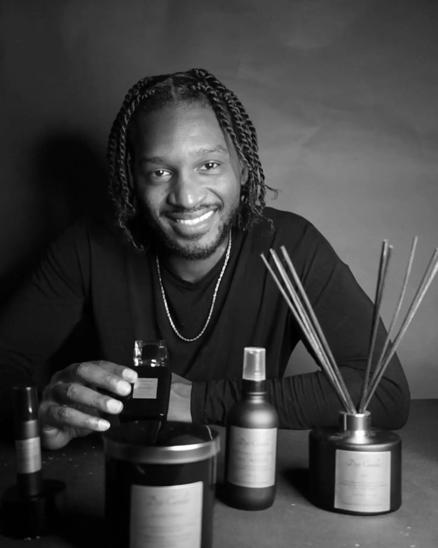 A smiling host in a black shirt poses with various fragrance products, including candles and room sprays, on a table.