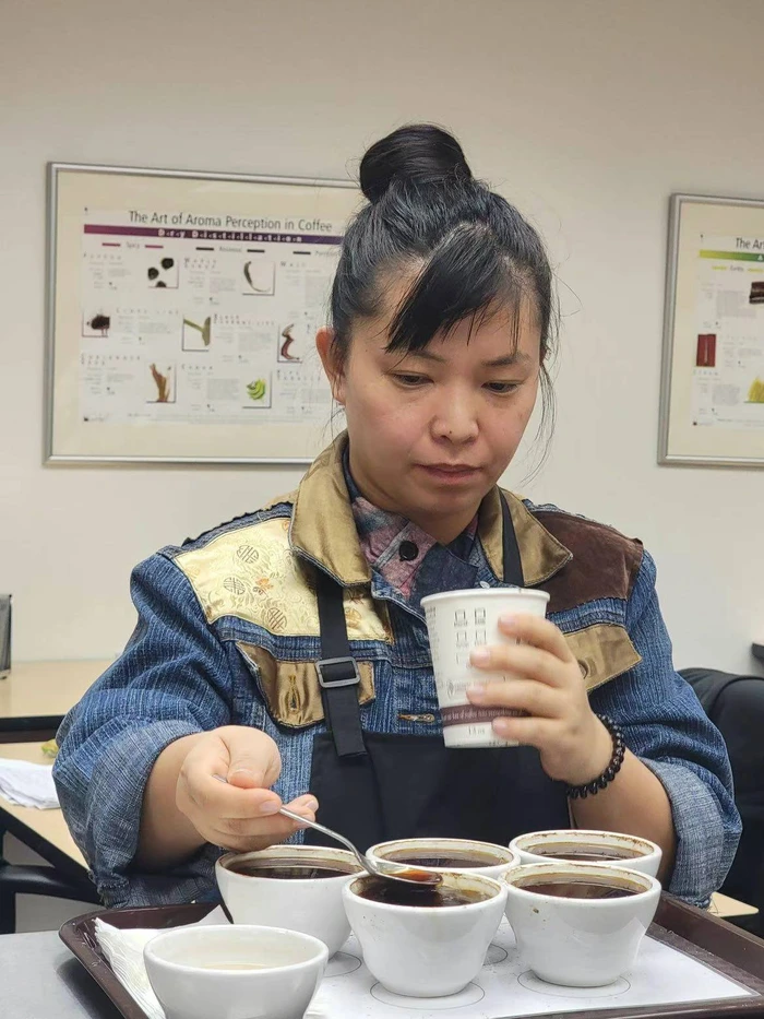 A barista examines coffee samples, holding a cup and spooning from bowls in a coffee tasting setup.