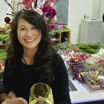 Smiling host with dark hair stands in front of a table filled with colorful flowers, ready for a terrarium workshop.