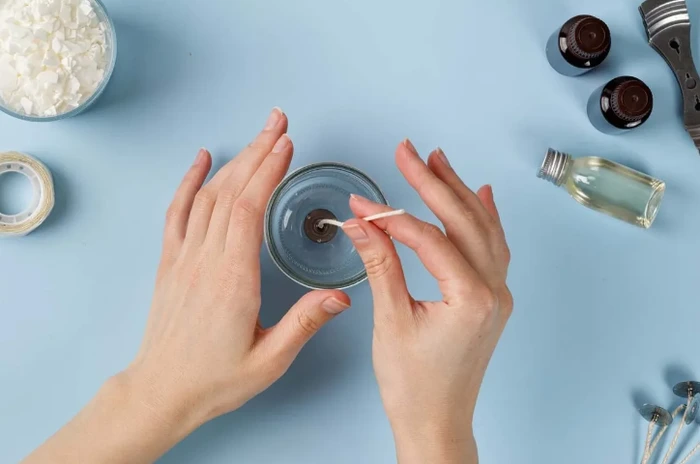 Hands carefully placing a wick in a clear candle container, with candle-making supplies on a light blue surface.