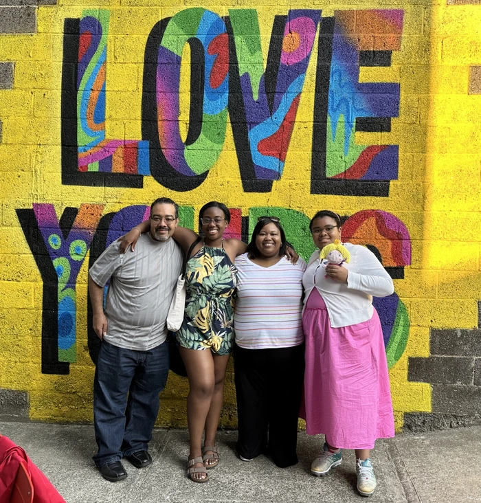 Group photo in front of a vibrant mural reading "LOVE YOURS," showcasing hosts of art classes and events.