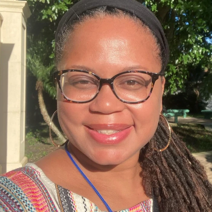 Smiling woman with glasses and dreadlocks against a sunny outdoor background, showcasing a vibrant, welcoming personality.
