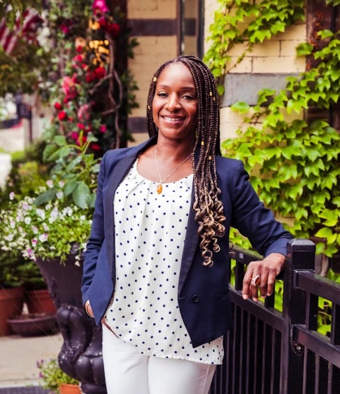 A smiling woman in a blazer and polka dot blouse stands against a backdrop of greenery, showcasing a welcoming vibe.