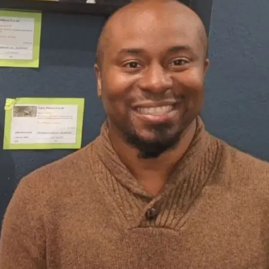 Smiling host in a brown sweater stands in front of art displays, promoting beauty through fine art.