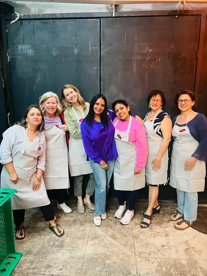 A smiling group of seven women in aprons poses together in a culinary workshop setting, ready for an engaging activity.