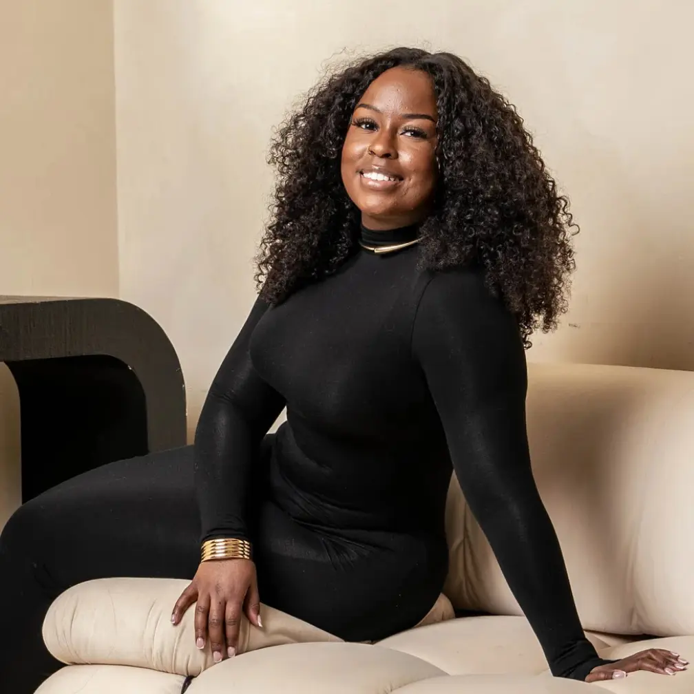 DeAndra Craigman, a fine artist and entrepreneur, smiles while seated in a stylish interior setting.