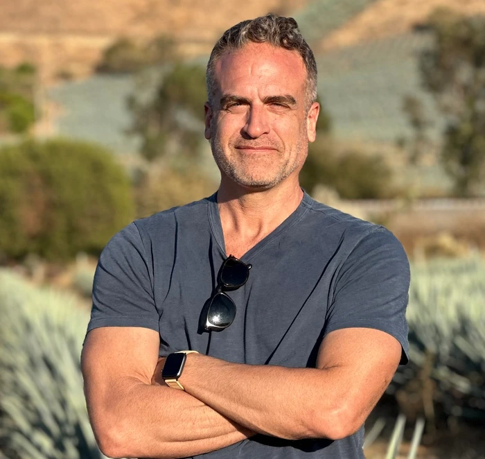 A smiling man stands outdoors with arms crossed, wearing a navy shirt and sunglasses, surrounded by greenery.
