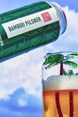 A can of Bamboo Pilsner is poured into a glass with a small palm tree decoration against a blue sky backdrop.