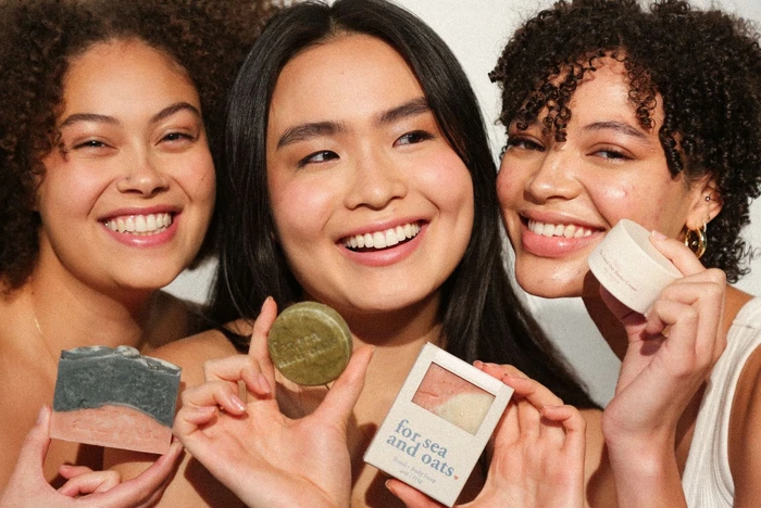 Three women smile while holding oat-based, zero-waste skincare products, showcasing their commitment to sensitive skin.