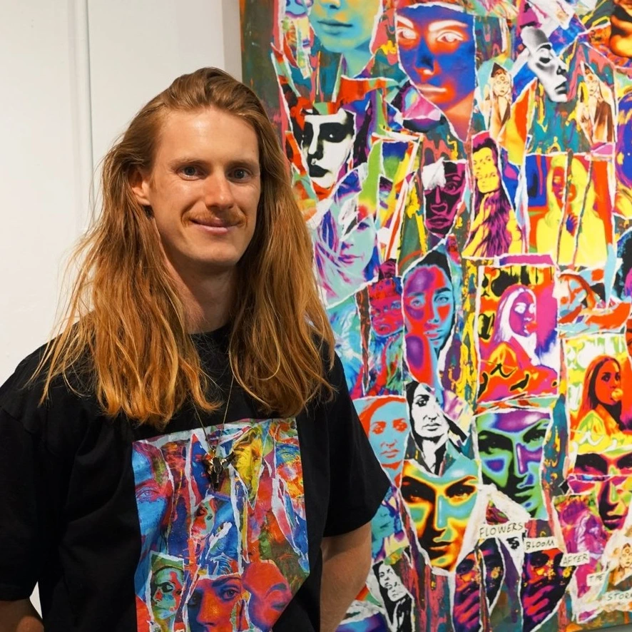 A smiling man with long hair stands in front of a colorful collage artwork, showcasing vibrant art and creativity.