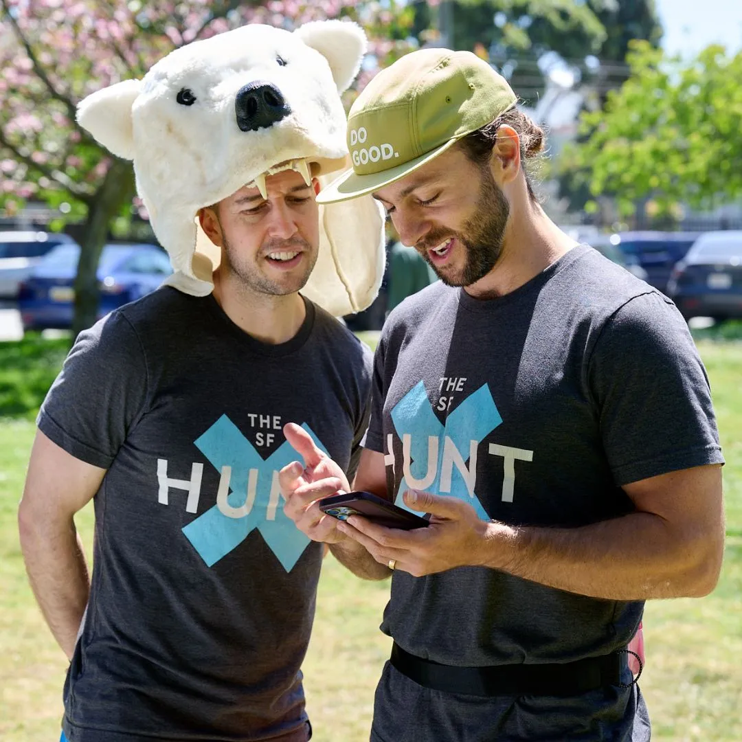 Two friends in fun hats hold a phone outdoors, sharing a moment in matching "The SF Hunt" shirts.
