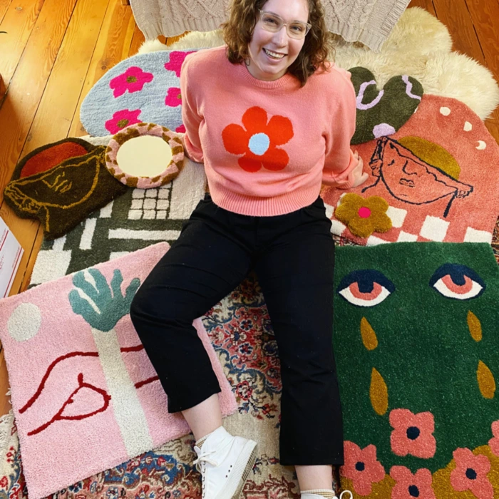 Hannah, a print and fiber artist, sits on colorful tufted rugs, smiling and showcasing her creative work in Queens, NY.