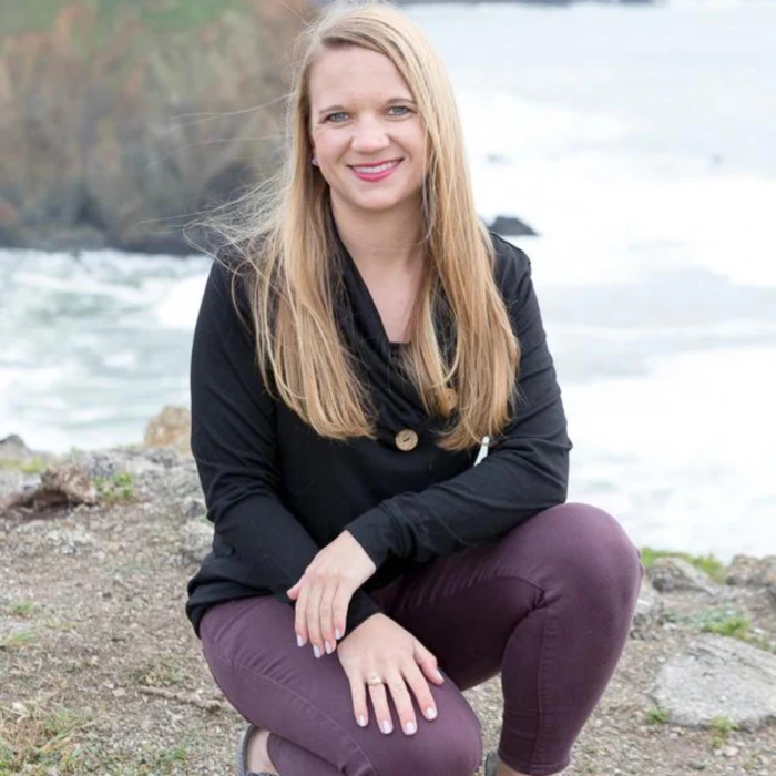 A smiling woman with long hair sits on a rocky coastline, showcasing her plant-based nail products.