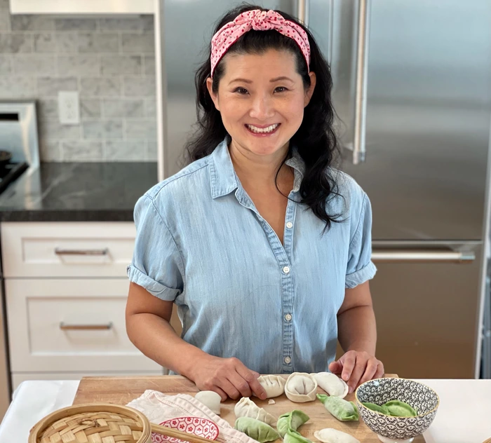 Judy Kim smiles while showcasing homemade dumplings in a kitchen, blending her culinary heritage and passion for cooking.