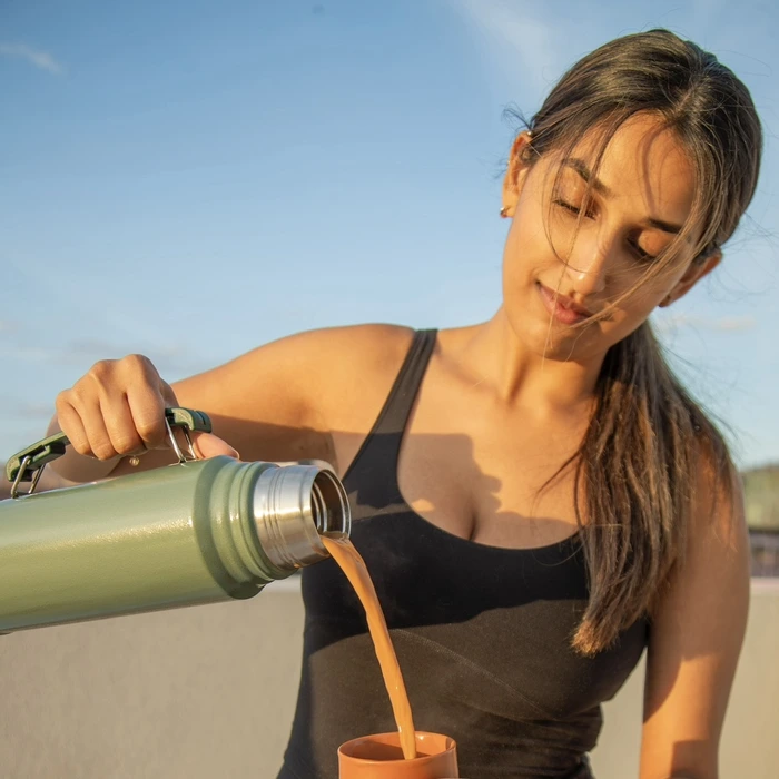 A woman pours chai from a thermos into a terracotta cup against a bright sky backdrop.