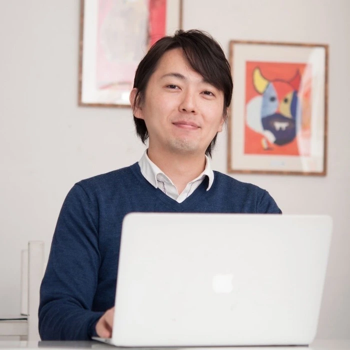 A smiling host in a blue sweater sits at a desk with a laptop, surrounded by colorful artwork.