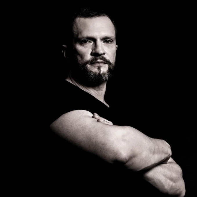 Portrait of a man with a beard and strong features, wearing a black shirt, arms crossed against a dark background.