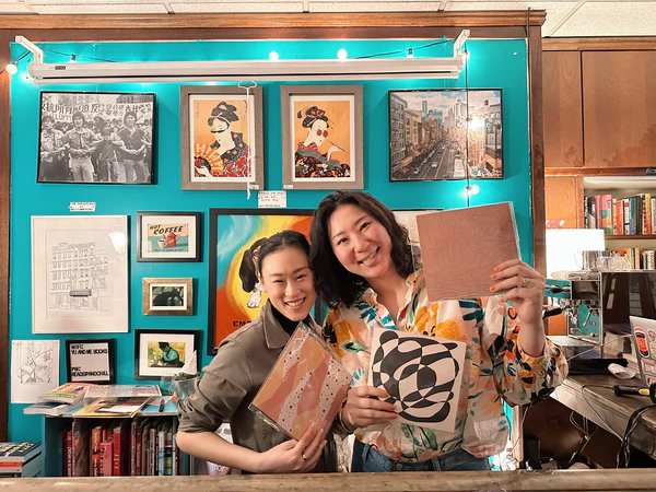 Two smiling hosts hold colorful stationery in a vibrant, art-filled shop, showcasing the creativity of handwritten letters.