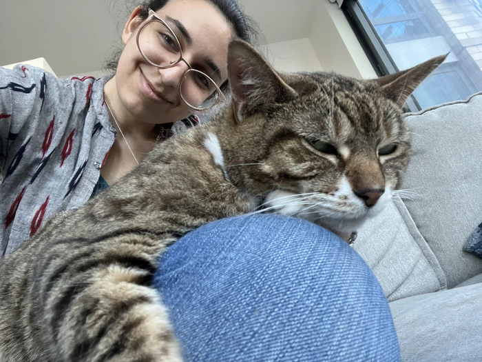A smiling woman with glasses poses with a sleeping tabby cat on her lap, sitting on a couch.