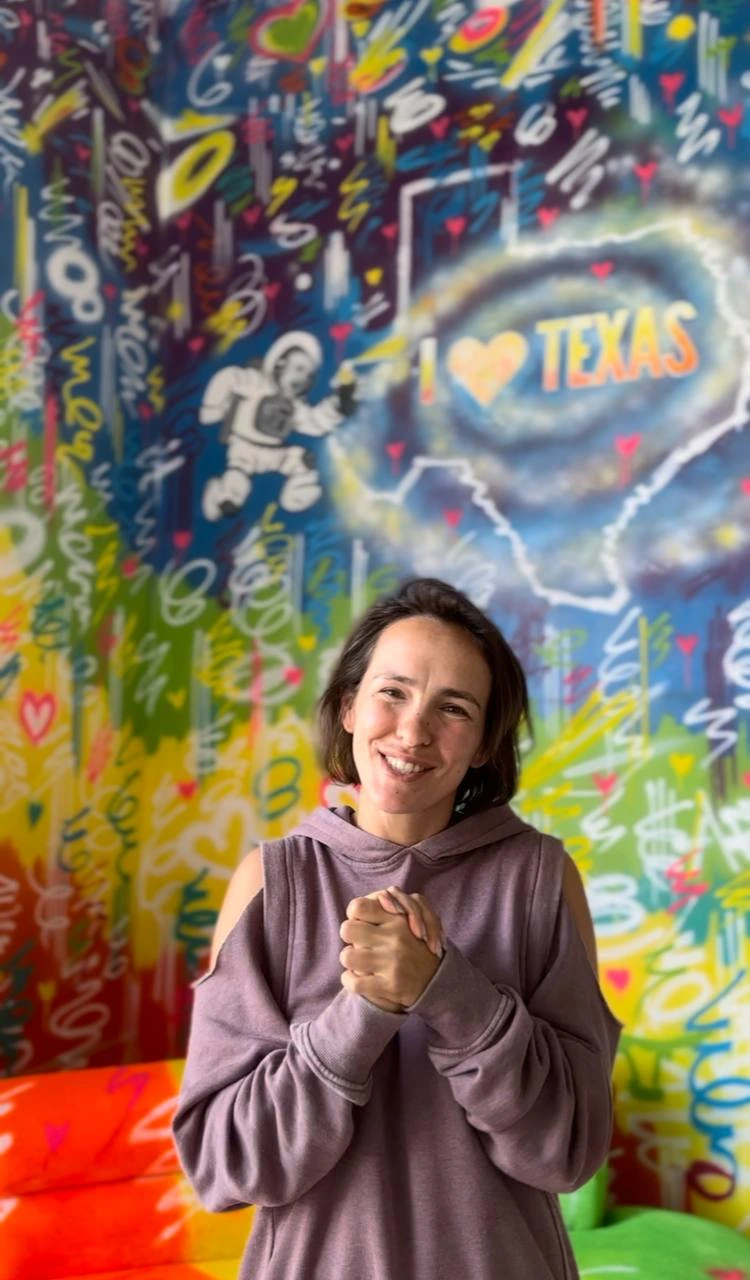Nadia smiles warmly in a colorful, graffiti-filled studio, embodying creativity and the spirit of art exploration.