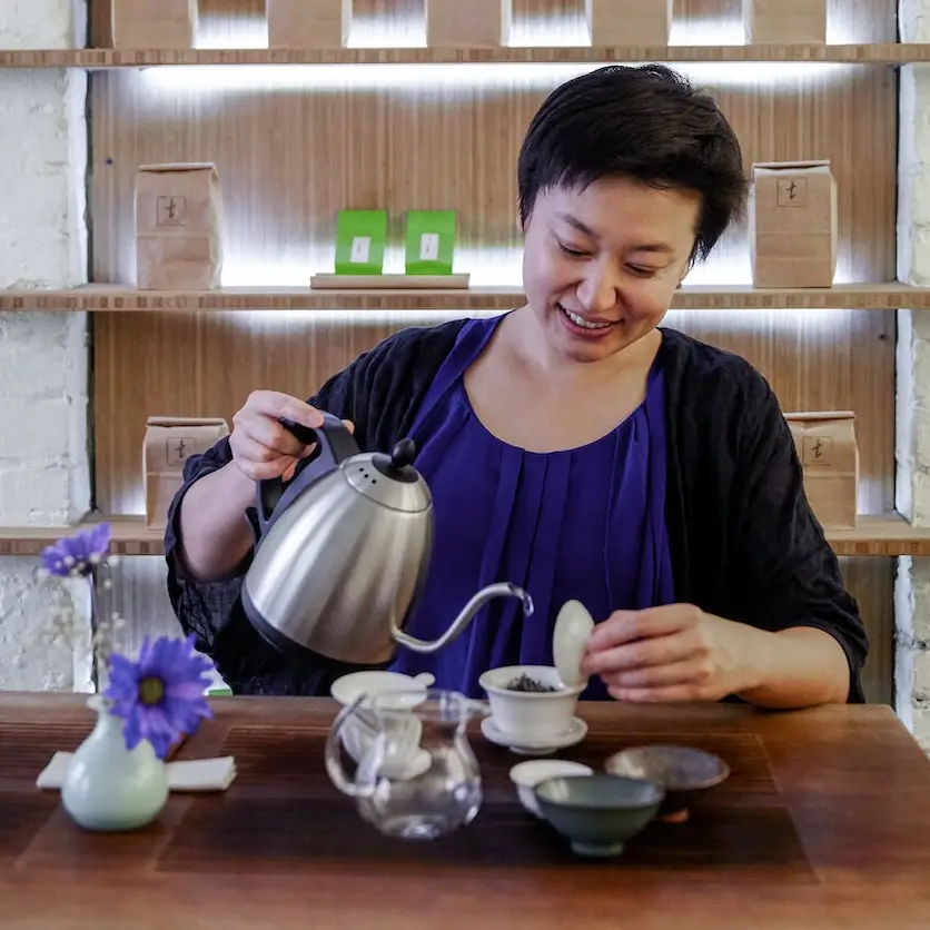 Shunan Teng pours tea, smiling, with tea tools and flowers on the table in a cozy tea shop setting.