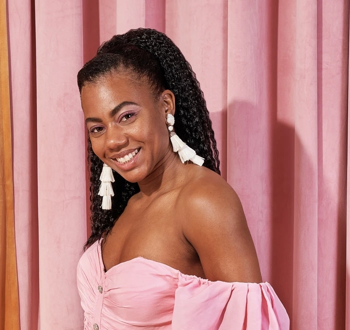 A smiling woman in a pink dress stands against pink curtains, promoting wellness and body care products and workshops.