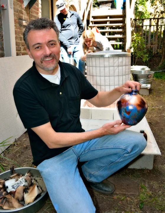 Steve Allen holds a colorful pottery piece, smiling in a workshop setting with people in the background.
