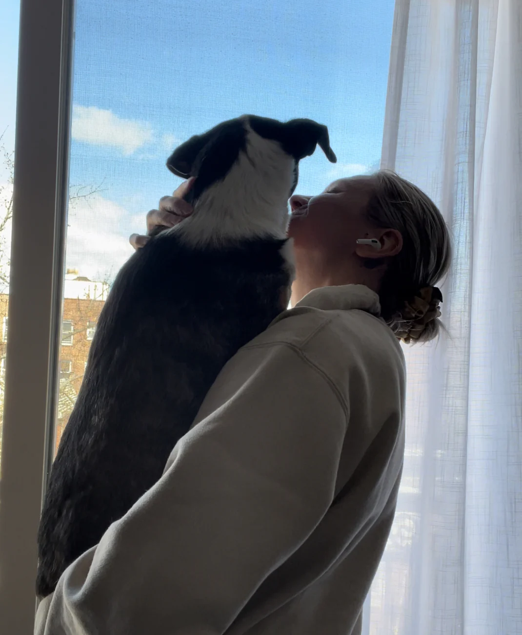 Emma holds her dog by a window, sharing a tender moment with blue skies and curtains in the background.