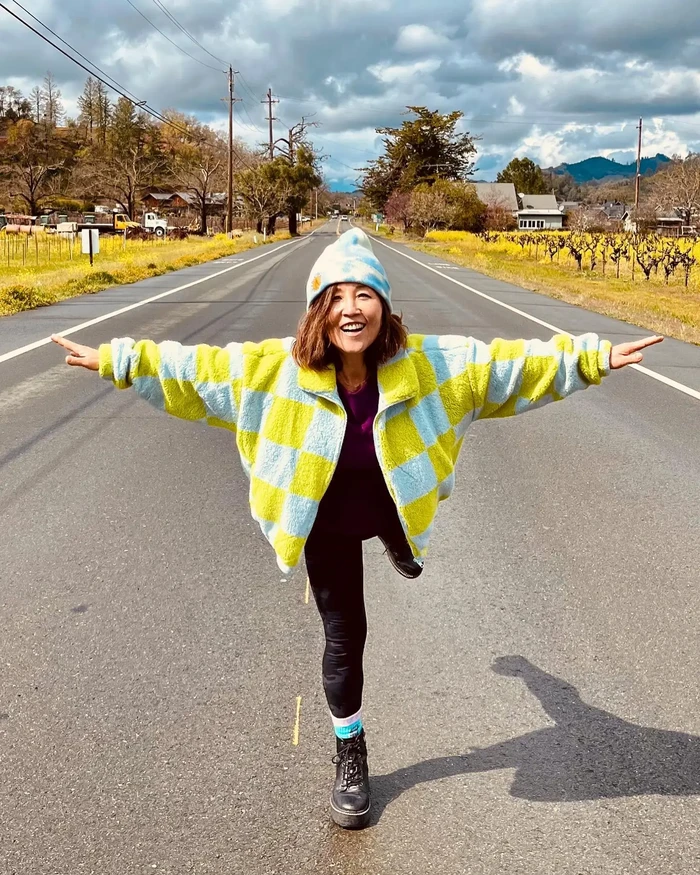 A joyful woman in a colorful jacket and beanie strikes a pose on a rural road surrounded by fields and trees.