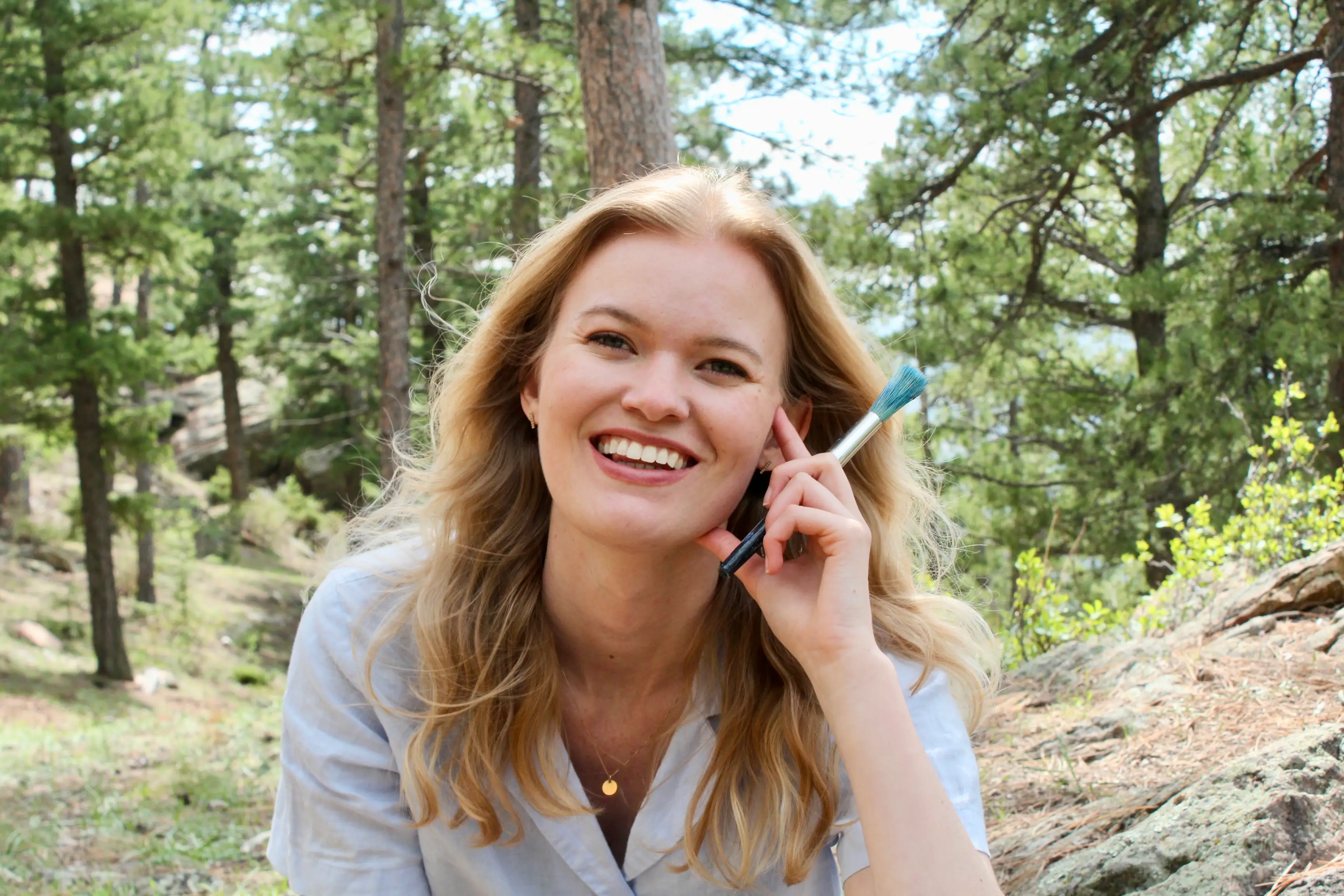 A smiling woman in a forest holds a paintbrush, promoting outdoor art experiences amidst nature.